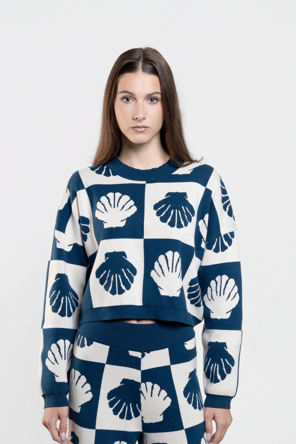 A woman stands against a light gray studio background wearing a matching navy and white knit set with a repeating shell pattern, consisting of a cropped long-sleeve top and wide-leg pants.