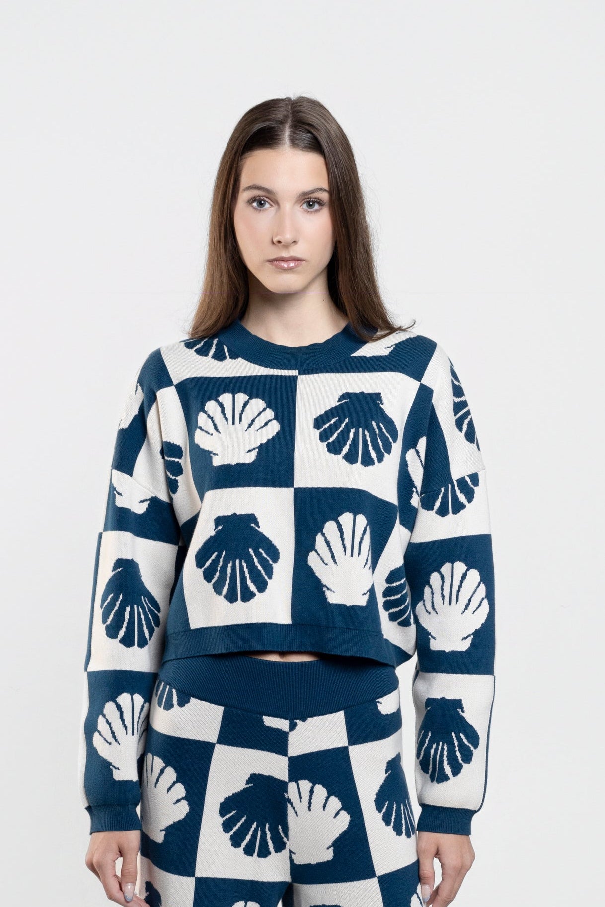 A woman stands against a light gray studio background wearing a matching navy and white knit set with a repeating shell pattern, consisting of a cropped long-sleeve top and wide-leg pants.