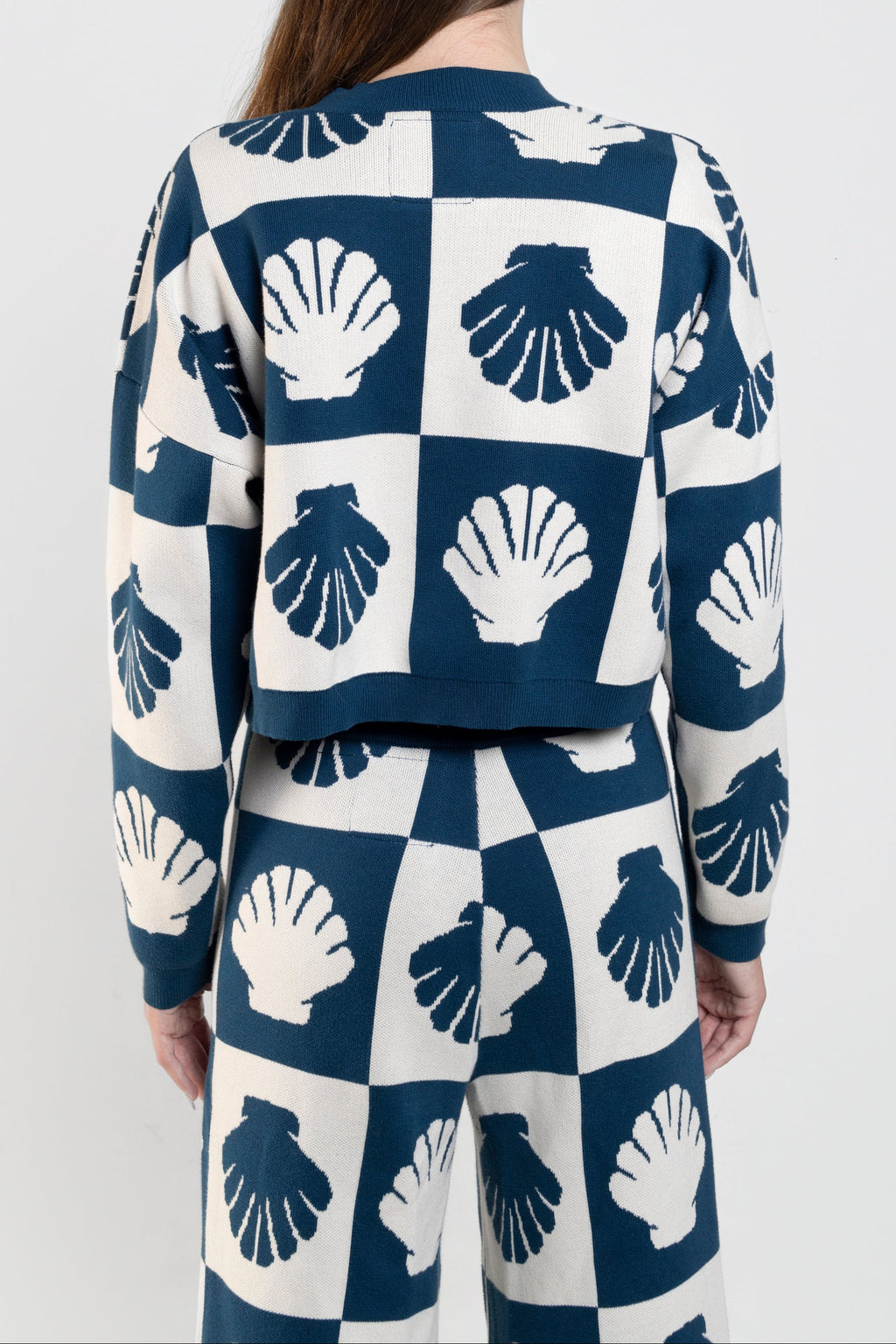 A model viewed from behind wearing a matching navy and white knit set with a repeating seashell pattern on a checkerboard background, against a plain light gray studio backdrop.