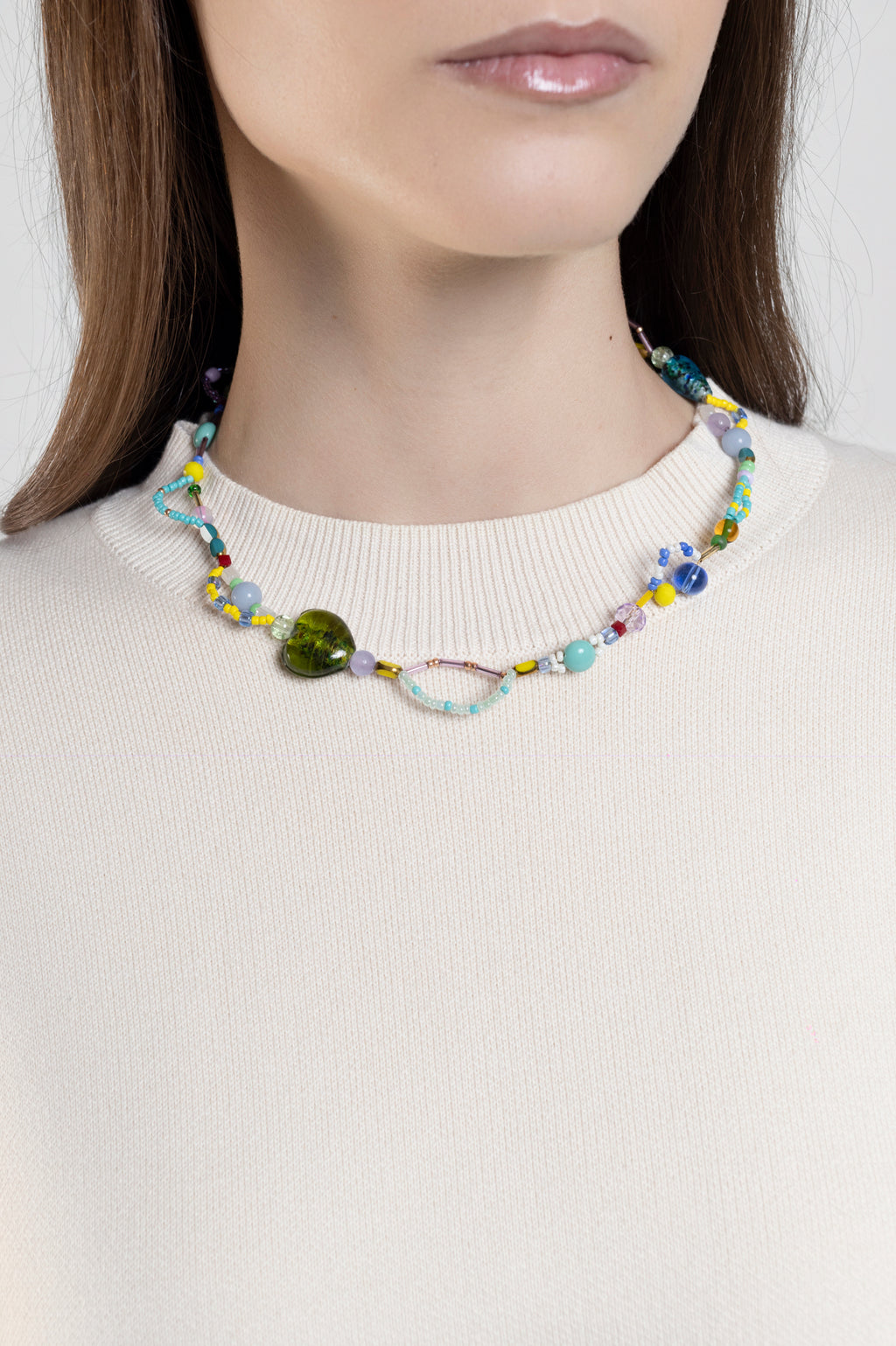 A close-up of a model wearing a colorful beaded necklace with a prominent green heart-shaped bead and a small metallic oval charm, paired with a cream-colored ribbed knit top.