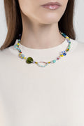 A close-up of a model wearing a colorful beaded necklace with a prominent green heart-shaped bead and a small metallic oval charm, paired with a cream-colored ribbed knit top.