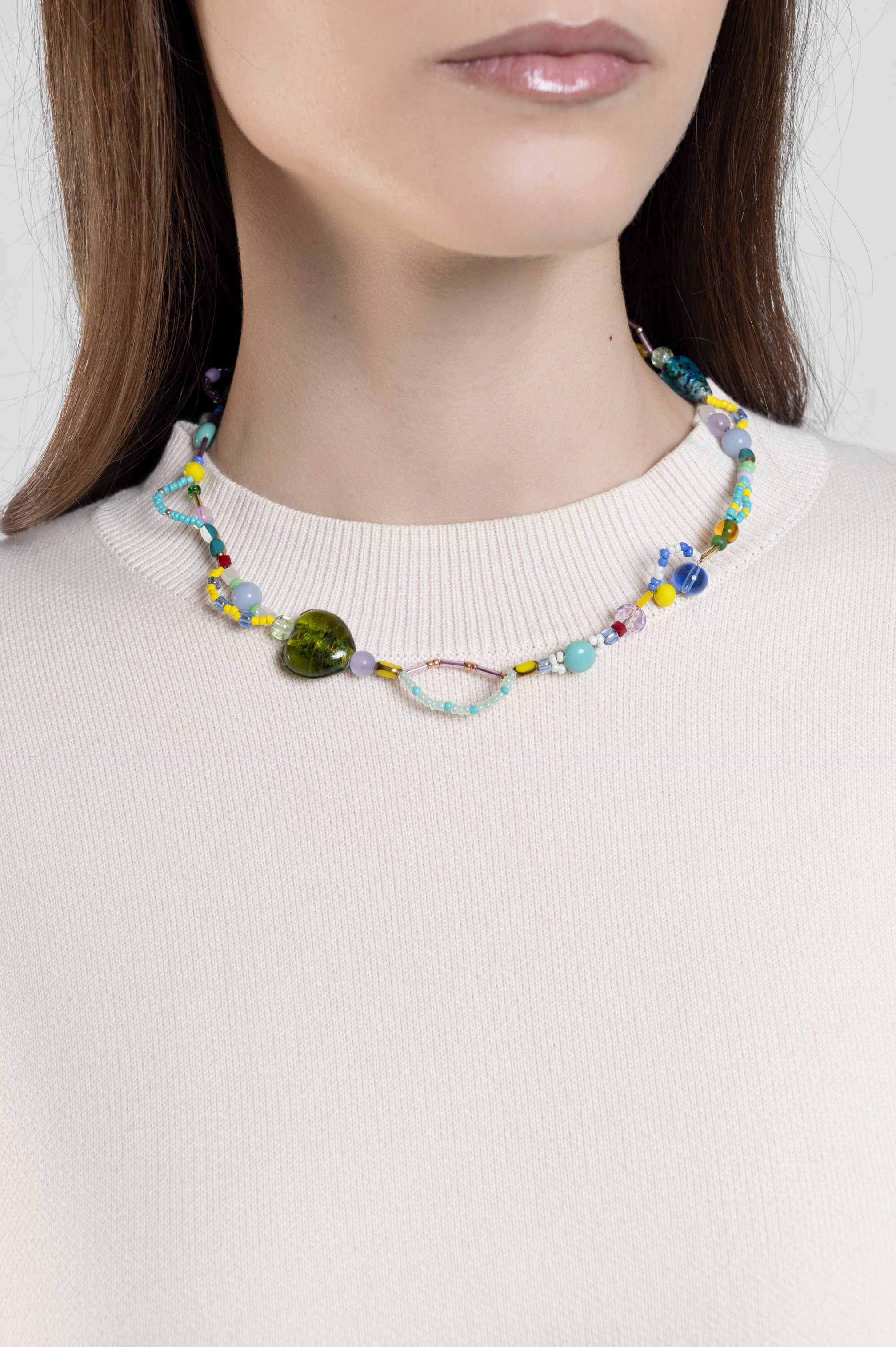 A close-up of a model wearing a colorful beaded necklace with a prominent green heart-shaped bead and a small metallic oval charm, paired with a cream-colored ribbed knit top.
