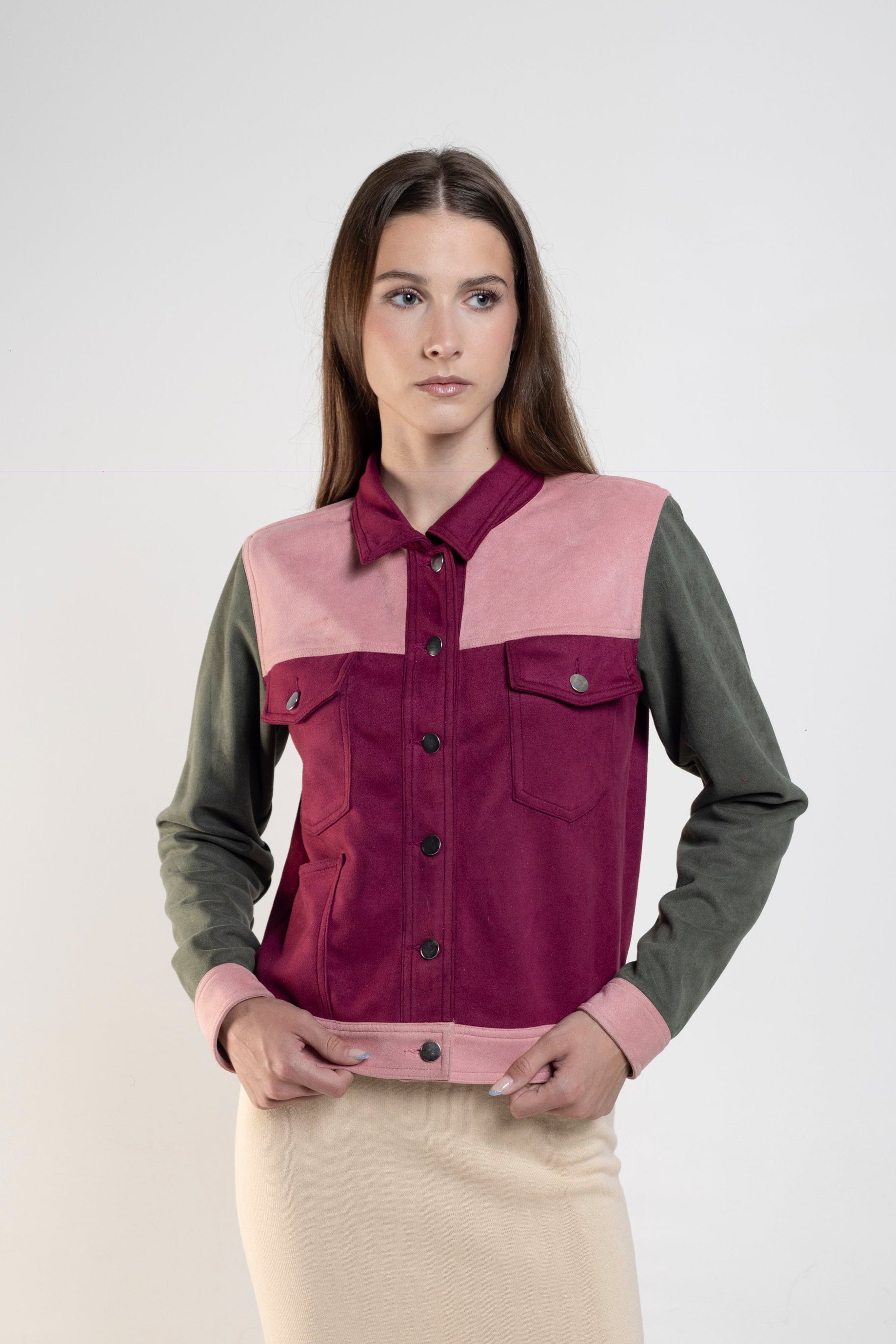 A woman modeling a color-blocked button-up shirt vest with a dusty pink yoke, deep magenta front panels, and olive green sleeves, paired with a cream-colored pencil skirt, against a plain light gray studio background.