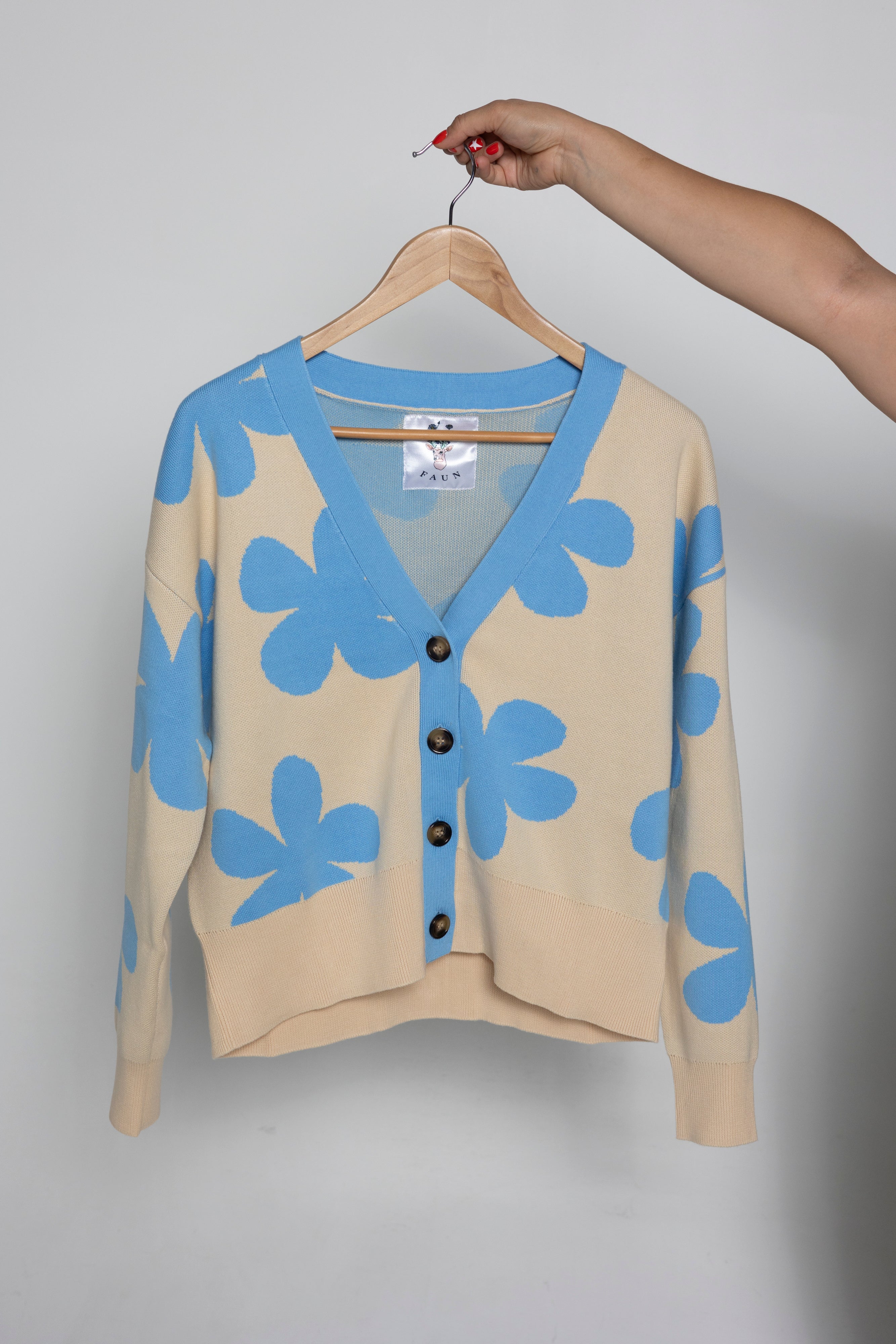 A beige cardigan sweater with large light blue floral motifs, hanging on a wooden hanger held by a person's hand against a plain light gray background.
