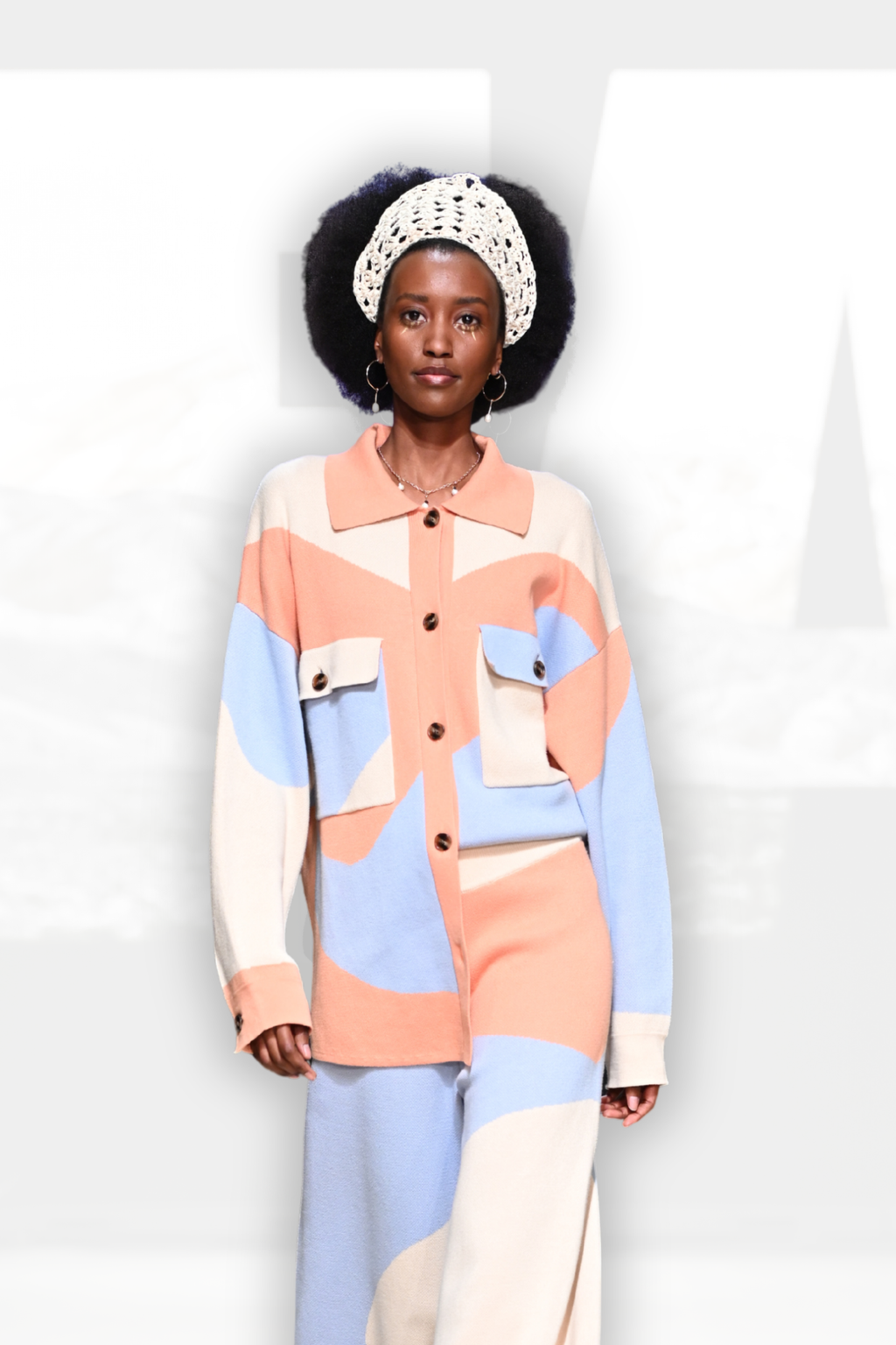A model wearing a matching color-blocked knit set consisting of a long-sleeve button-up shirt and wide-leg trousers. The color-blocked pattern features soft pastel shades of peach, light blue, cream, and beige.