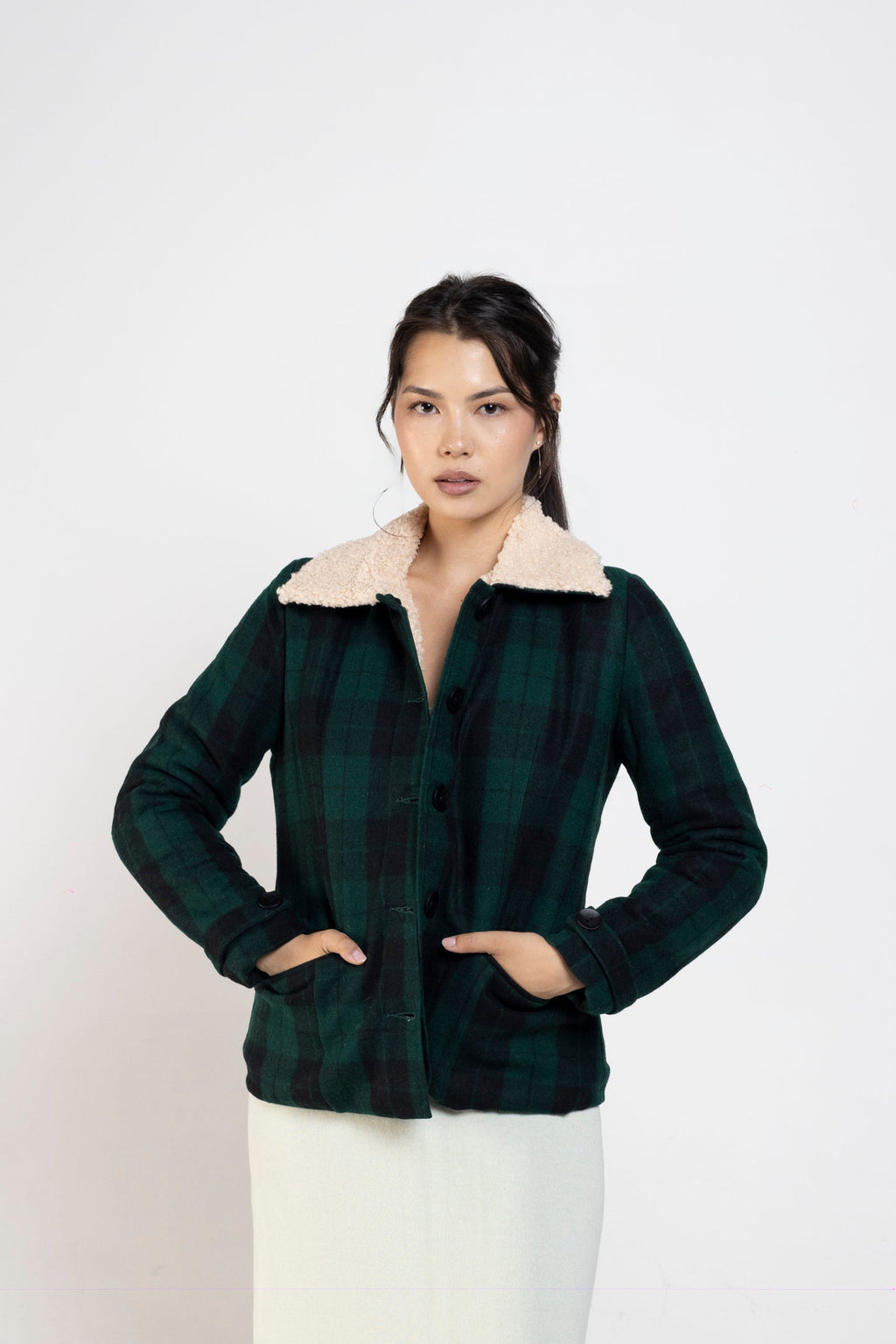 A woman modeling a dark green and black plaid cropped jacket with a cream-colored collar, paired with a long cream skirt, standing against a plain light gray studio background.
