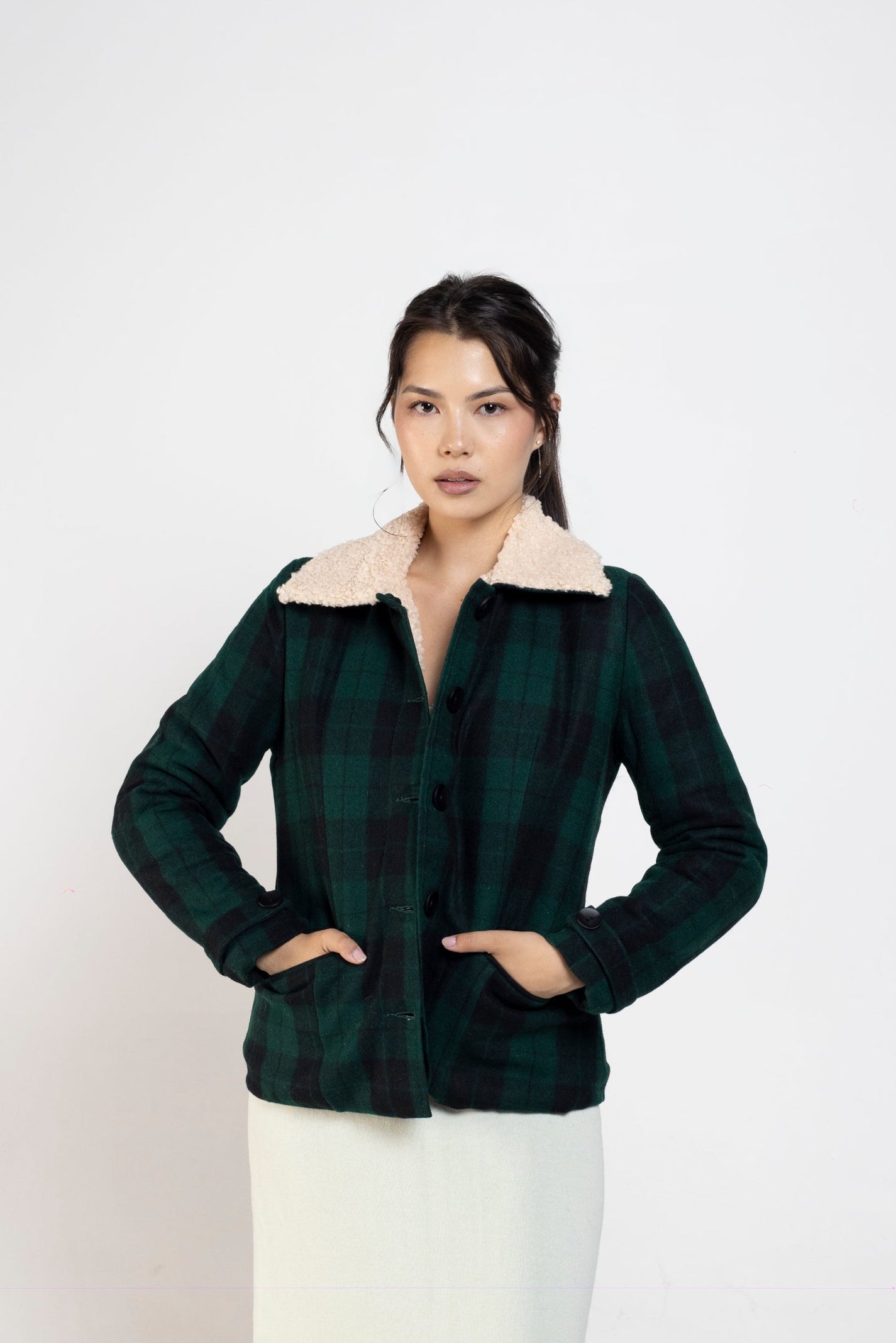 A woman modeling a dark green and black plaid cropped jacket with a cream-colored collar, paired with a long cream skirt, standing against a plain light gray studio background.