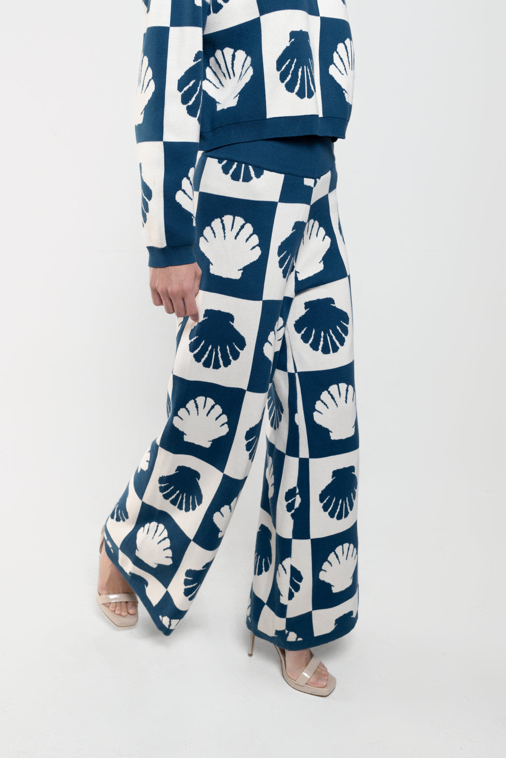 A model wearing a matching navy blue and white checkered set with a scallop shell pattern, featuring wide-leg trousers and a cropped long-sleeve top, paired with clear high-heeled sandals.