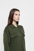 A woman modeling an olive green long-sleeve utility shirt dress with a pointed collar and front button closure, photographed against a plain light gray studio background.